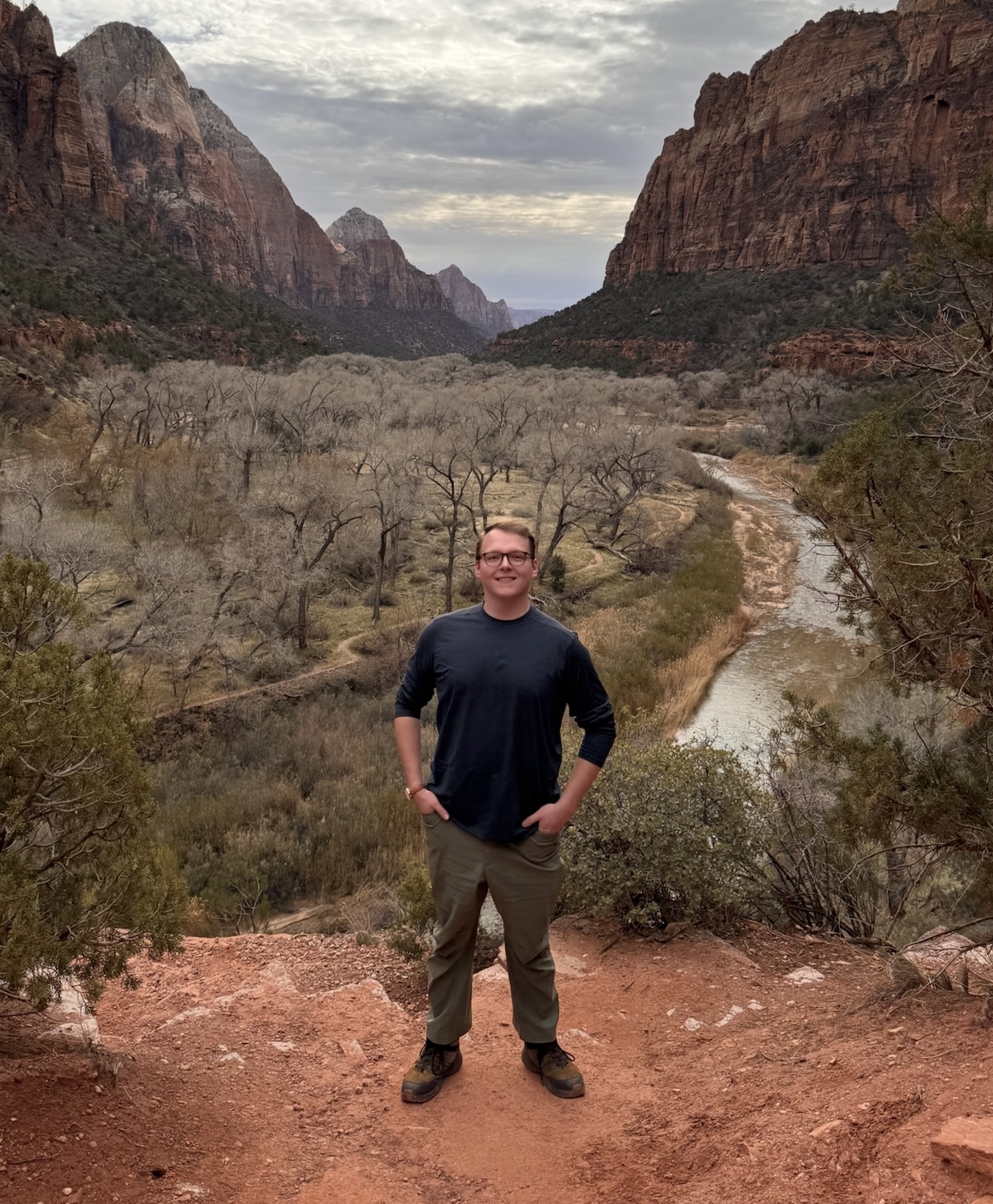 Dane standing on a cliff overlook in Zion National Park, with red canyon walls and the Virgin River below.